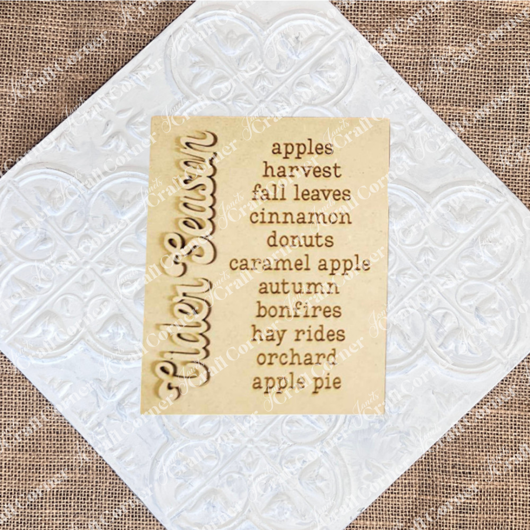The image beautifully showcases Janet's Craft Corner's "Cider Words Interchangeable Sign" DIY home decor craft kit. The wooden sign, engraved with "Cider Season," is complemented on the right by lively words that evoke apples, harvest, and fall leaves. Together, they conjure the cozy essence of autumn with cinnamon donuts, caramel apple treats, bonfires, hay rides at the orchard, and apple pie delights.