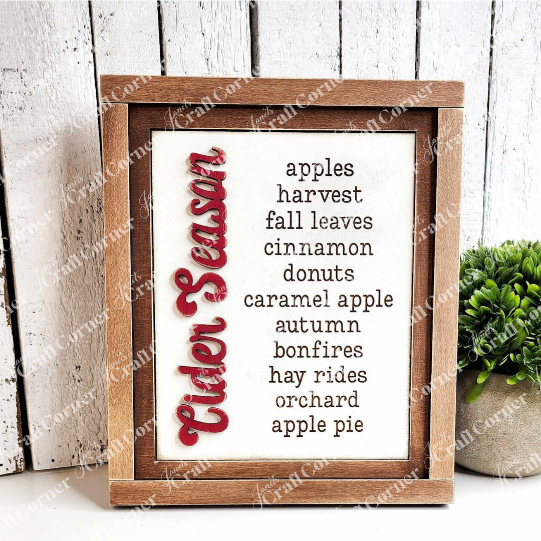 On the left, Janet's Craft Corner's Cider Words Interchangeable Sign, part of a DIY home decor craft kit, showcases "Cider Season" in vibrant red script. The right side displays cozy words such as "apples," "harvest," "fall leaves," and "cinnamon." A nearby small potted plant introduces a touch of nature to this charming craft insert scene.