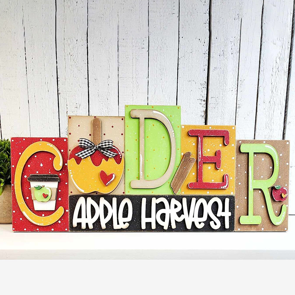 The Cider Word Block Shelf Sitter from Janet's Craft Corner spells "Coder" with decorative wood letters. Each letter is hand-painted for the fall season, featuring charming designs of apples and polka dots. Below the letters, a sign proudly displays the words "Apple Harvest" in playful white script. This delightful DIY home decor kit is perfect for adding a festive touch to your space against a white rustic wooden wall backdrop.