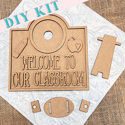 Janet's Craft Corner offers the Welcome to Our Classroom Interchangeable Set, featuring a wooden sign with "Welcome to Our Classroom" text on a textured background, along with apple and pencil cutouts and extra assembly pieces.