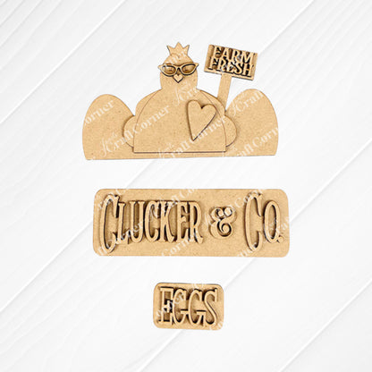 Wooden cutouts on a textured surface create a charming chicken motif. This decorative piece, featuring a chicken shape with glasses, a "Farm Fresh" sign, and an "EGGS" cutout, is part of the Clucker & Co. Interchangeable Insert DIY home decor kit by Janet's Craft Corner. The ensemble is beautifully displayed on a white wooden background.