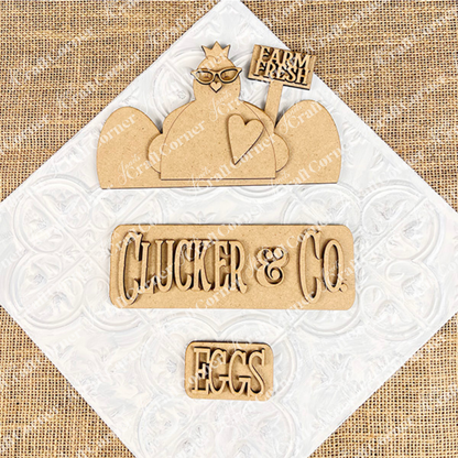 Wooden cutouts on a textured surface create a charming chicken motif. This decorative piece, featuring a chicken shape with glasses, a "Farm Fresh" sign, and an "EGGS" cutout, is part of the Clucker & Co. Interchangeable Insert DIY home decor kit by Janet's Craft Corner. The ensemble is beautifully displayed against a burlap background.