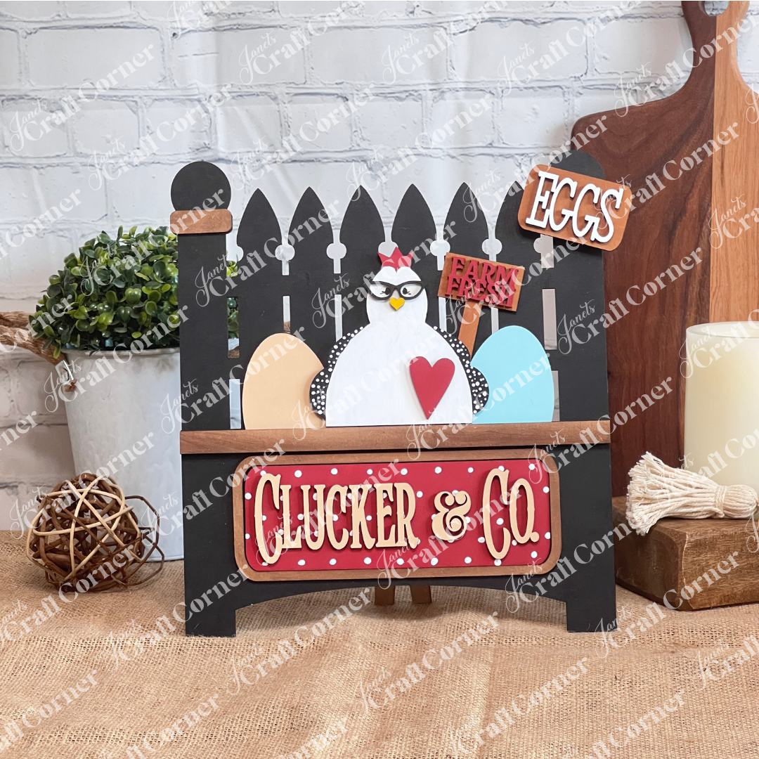 Janet's Craft Corner introduces the Clucker & Co. Interchangeable Insert - Painted, a decorative piece featuring a delightful chicken motif adorned with glasses, alongside a red heart and colorful eggs. The sign displays "EGGS" and "Farm Fresh" at the top, with "Clucker & Co." beneath on a black garden fence, making it an ideal choice for farmhouse decor enthusiasts seeking versatile craft inserts.