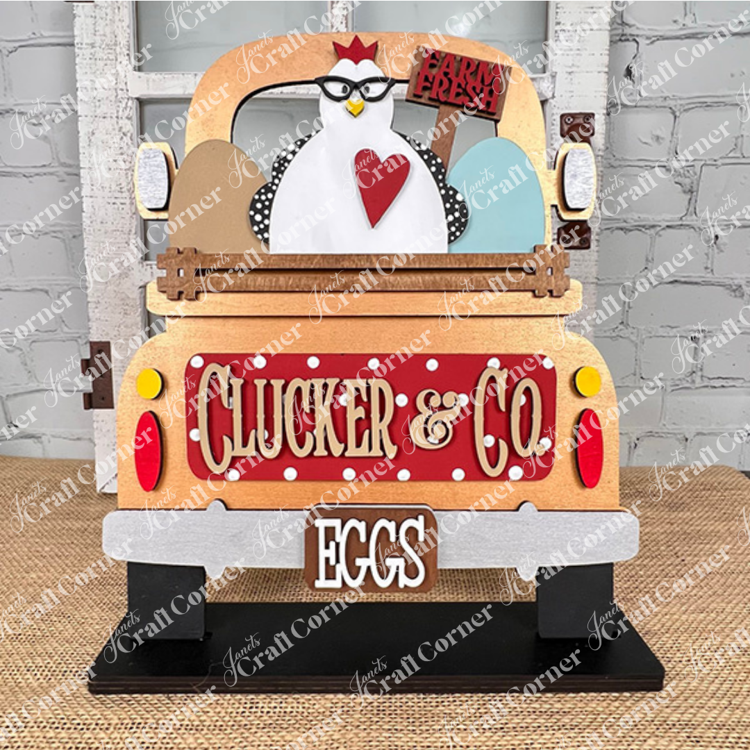 A rustic DIY home decor kit from Janet's Craft Corner showcases a chicken atop an antique truck base labeled "Clucker & Co. Eggs," holding a "Farm Fresh" sign, with three eggs nestled in the back, all set against a textured surface and brick wall. Ideal for fans of hand-painted crafts and rustic charm.