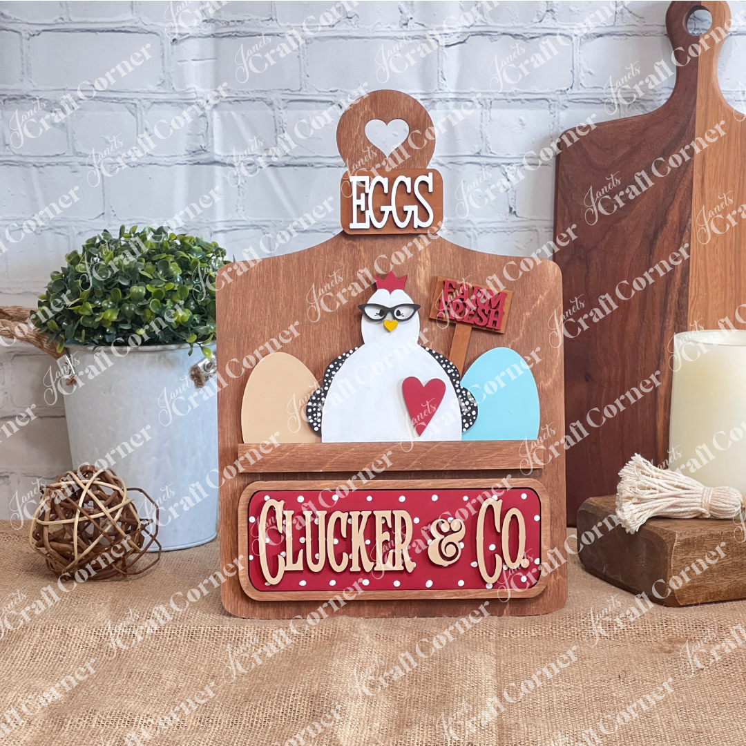 Janet's Craft Corner offers a charming DIY kit with a decorative breadboard featuring a cartoon chicken, eggs, and "Farm Fresh" sign. It reads "EGGS" on top and "CLUCKER & CO." below, with interchangeable heart and bow accessories against a rustic backdrop.