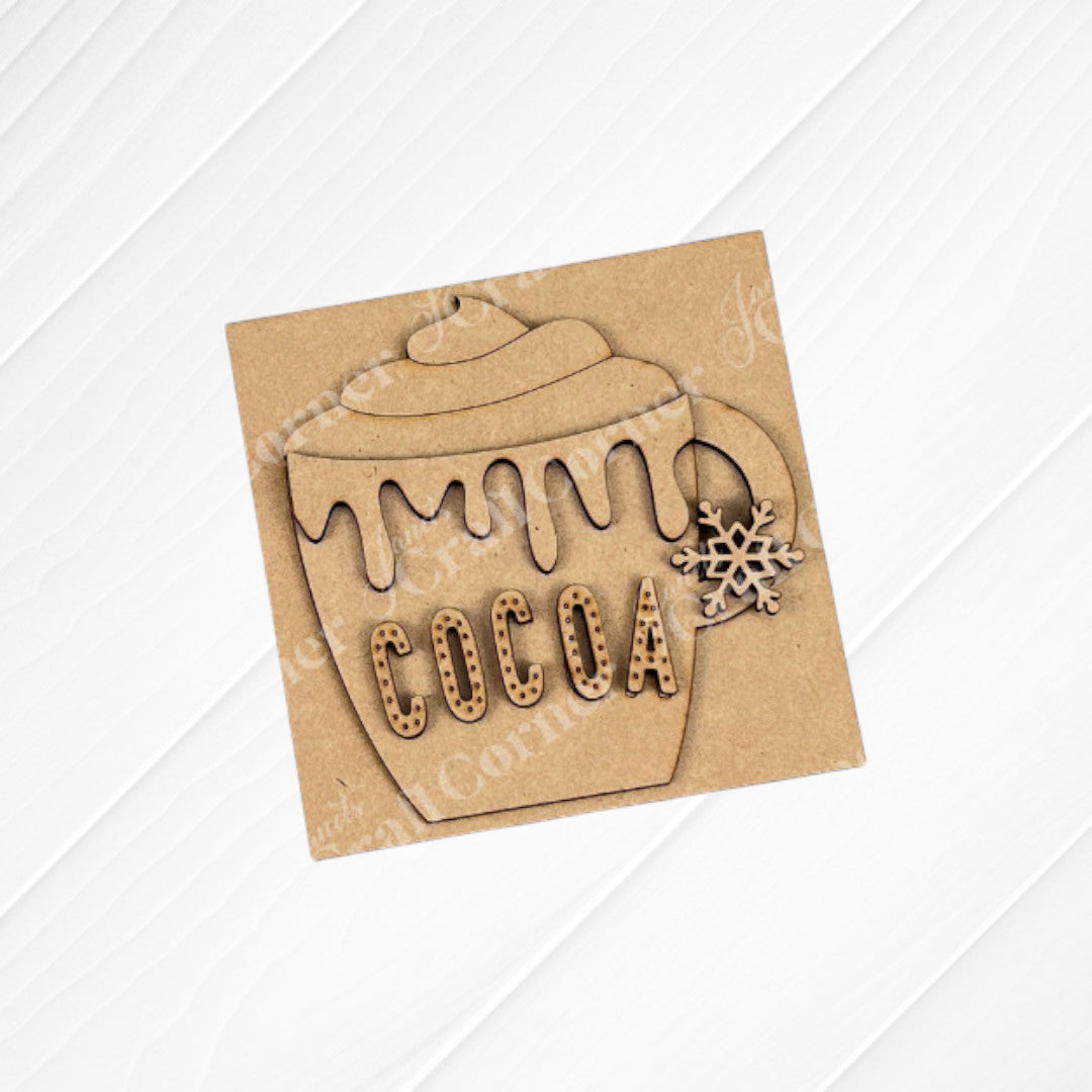 The Cup of Cheer Hot Cocoa Mini Shelf Sitter by Janet's Craft Corner is a wooden cocoa mug cutout with "COCOA" and a snowflake—ideal as DIY seasonal home decor. Shown on a brown square with a white wood background. Set of 1.