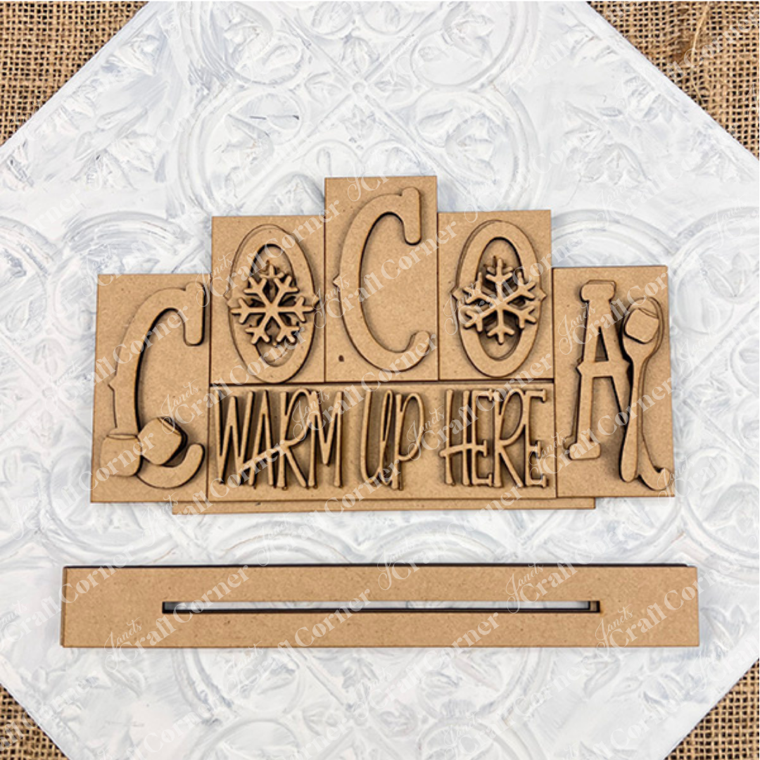 The Cocoa Mini Word Block by Janet's Craft Corner features "COCOA WARM UP HERE" with snowflakes, a mitten, and a mug on a textured white background. This hand-painted wooden sign is perfect for cozy home décor.