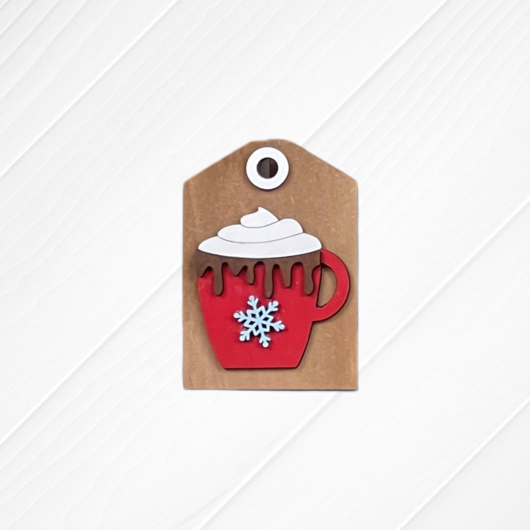 The Cup of Cheer Hot Cocoa Mini Shelf Sitters by Janet's Craft Corner features a brown gift tag with a red mug, whipped cream, chocolate drips, and blue snowflake—perfect hot cocoa bar decor on a white textured background.