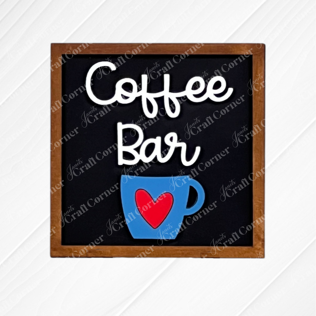 A small sign reading "Coffee Bar" with a blue coffee cup and red heart sits on a stand—perfect inspiration for your next Coffee Bar Swappable Squares DIY kit from Janet’s Craft Corner. Displayed on a white wood background.