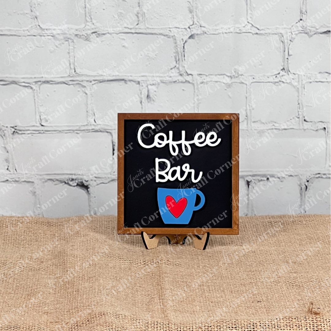 A small sign reading "Coffee Bar" with a blue coffee cup and red heart sits on a stand—perfect inspiration for your next Coffee Bar Swappable Squares DIY kit from Janet’s Craft Corner. Display in front of brick or burlap for cozy charm!.