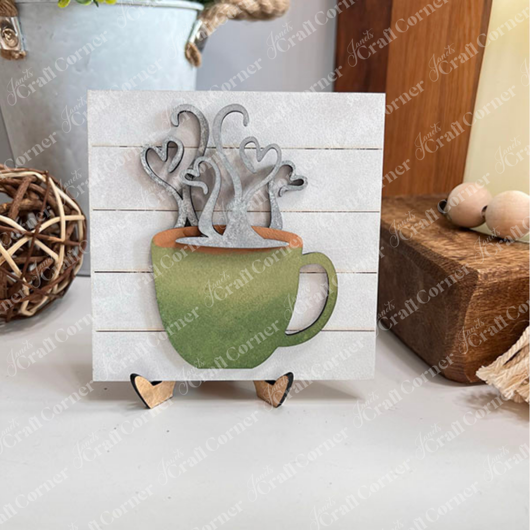 Janet's Craft Corner Coffee Swappable Squares is a DIY seasonal home decor craft kit featuring a wooden coffee mug cutout with heart steam, set in a farmhouse decor setting. With a green cup and brown "coffee" it is ideal for coffee lovers and creative projects. 