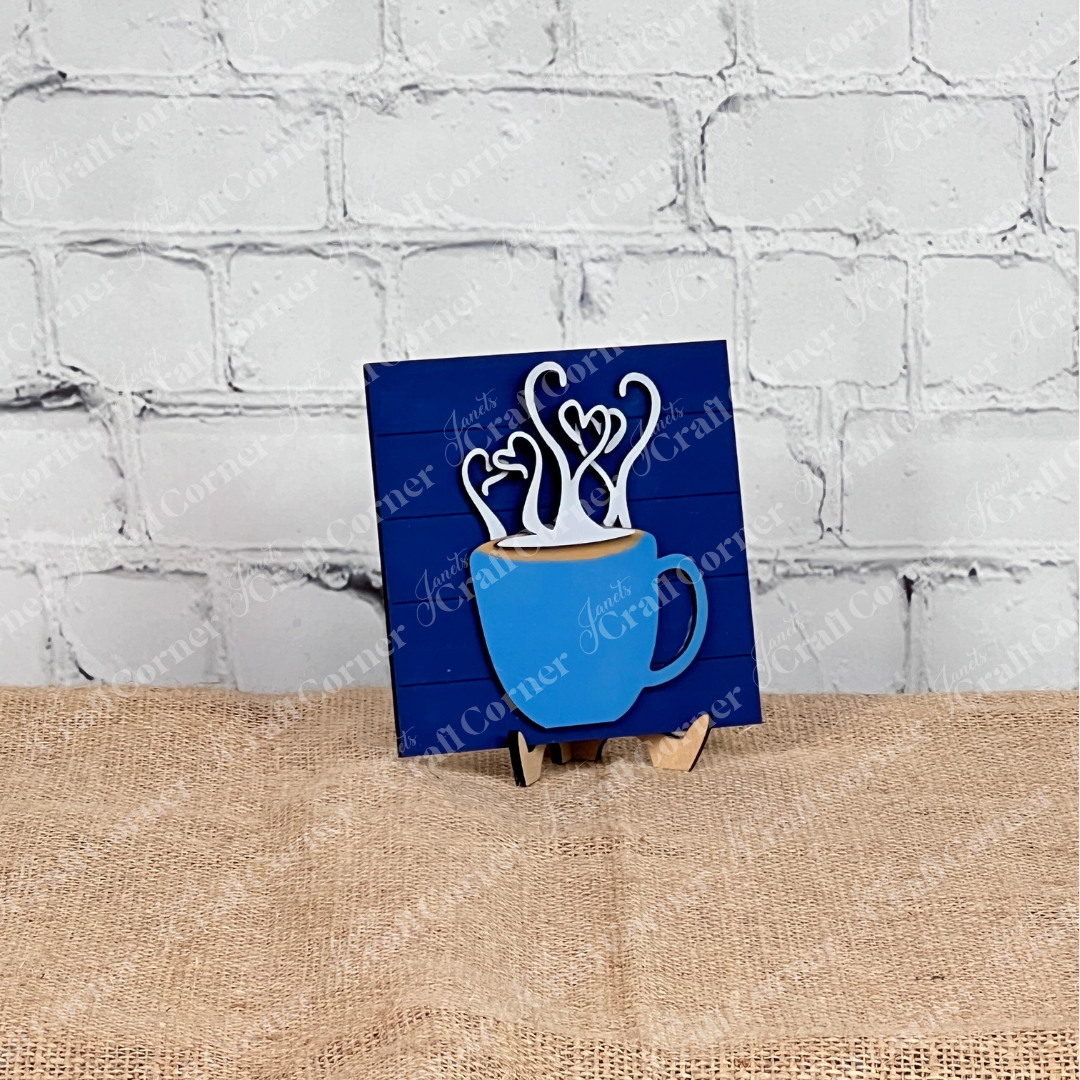 Janet's Craft Corner Coffee Swappable Squares is a DIY seasonal home decor craft kit featuring a wooden coffee mug cutout with heart steam, set in front of a white brick background. With a blue cup and brown "coffee" it is ideal for coffee lovers and creative projects. 