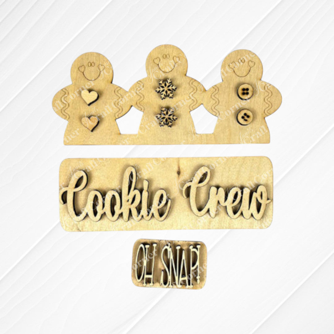 The Cookie Crew Interchangeable Insert by Janet's Craft Corner is an ideal DIY home decor kit, featuring wooden cutouts of three gingerbread men adorned with heart, snowflake, and button designs. A sign at the base displays "Cookie Crew" with a smaller "Oh Snap!" text, all crafted from light-colored wood.