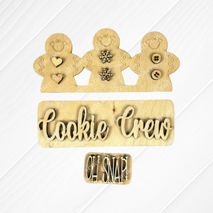 The Cookie Crew Interchangeable Insert by Janet's Craft Corner is an ideal DIY home decor kit, featuring wooden cutouts of three gingerbread men adorned with heart, snowflake, and button designs. A sign at the base displays "Cookie Crew" with a smaller "Oh Snap!" text, all crafted from light-colored wood.
