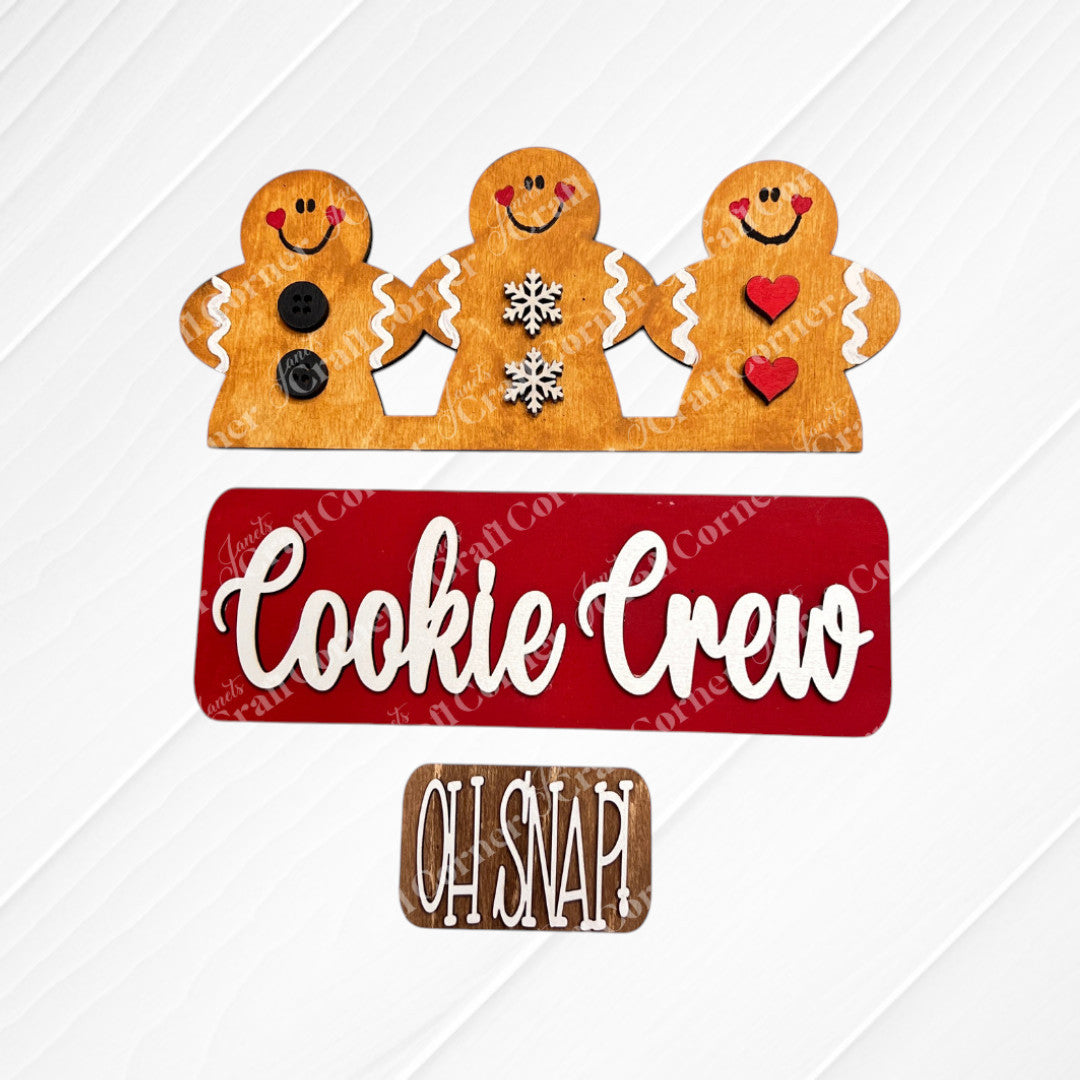 Three cheerful gingerbread figures—one with buttons, another with snowflakes, and the last with hearts—embellish a red sign that says "Cookie Crew," accompanied by a brown sign that reads "Oh Snap!" on a gray background. This charming DIY home decor kit from Janet's Craft Corner provides interchangeable decorations to brighten up the festive season.