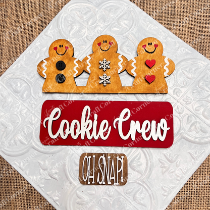 Three cheerful gingerbread figures—one with buttons, another with snowflakes, and the last with hearts—embellish a red sign that says "Cookie Crew," accompanied by a brown sign that reads "Oh Snap!" on a gray background. This charming DIY home decor kit from Janet's Craft Corner provides interchangeable decorations to brighten up the festive season.