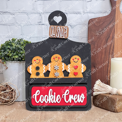 The Cookie Crew Interchangeable Insert by Janet's Craft Corner is a DIY home decor craft kit featuring three uniquely patterned gingerbread men on a white picket fence. The board reads "Oh Snap!" and includes a red "Cookie Crew" sign against a white brick wall, perfect for seasonal inserts.