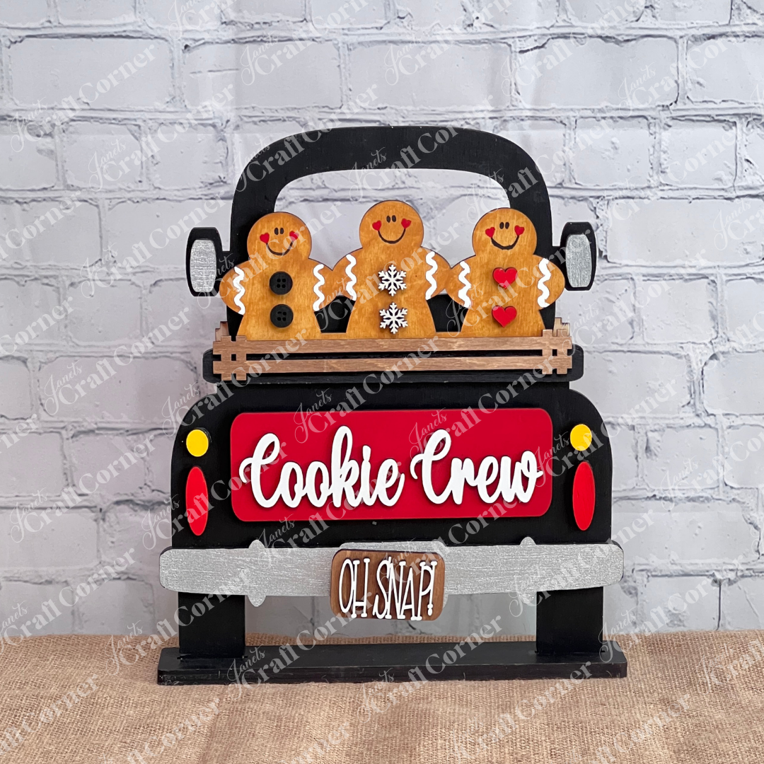A delightful festive decoration from Janet's Craft Corner highlights three cheerful gingerbread figures riding in an antique truck labeled "Cookie Crew." Below, a small wooden sign reads "Oh Snap!" The truck, featuring white and brown accents, serves as the charming base for customizable inserts, offering a personalized touch. This is part of the Antique Truck Base with Interchangeable Insert DIY home decor kit.