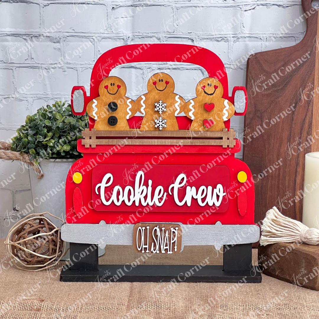 The Cookie Crew Interchangeable Insert from Janet's Craft Corner showcases three delightful gingerbread figures riding in a red truck labeled "Cookie Crew," with the whimsical phrase "Oh Snap!" underneath. Perfect as versatile decor, these gingerbread men are beautifully detailed with buttons, a snowflake, and hearts.