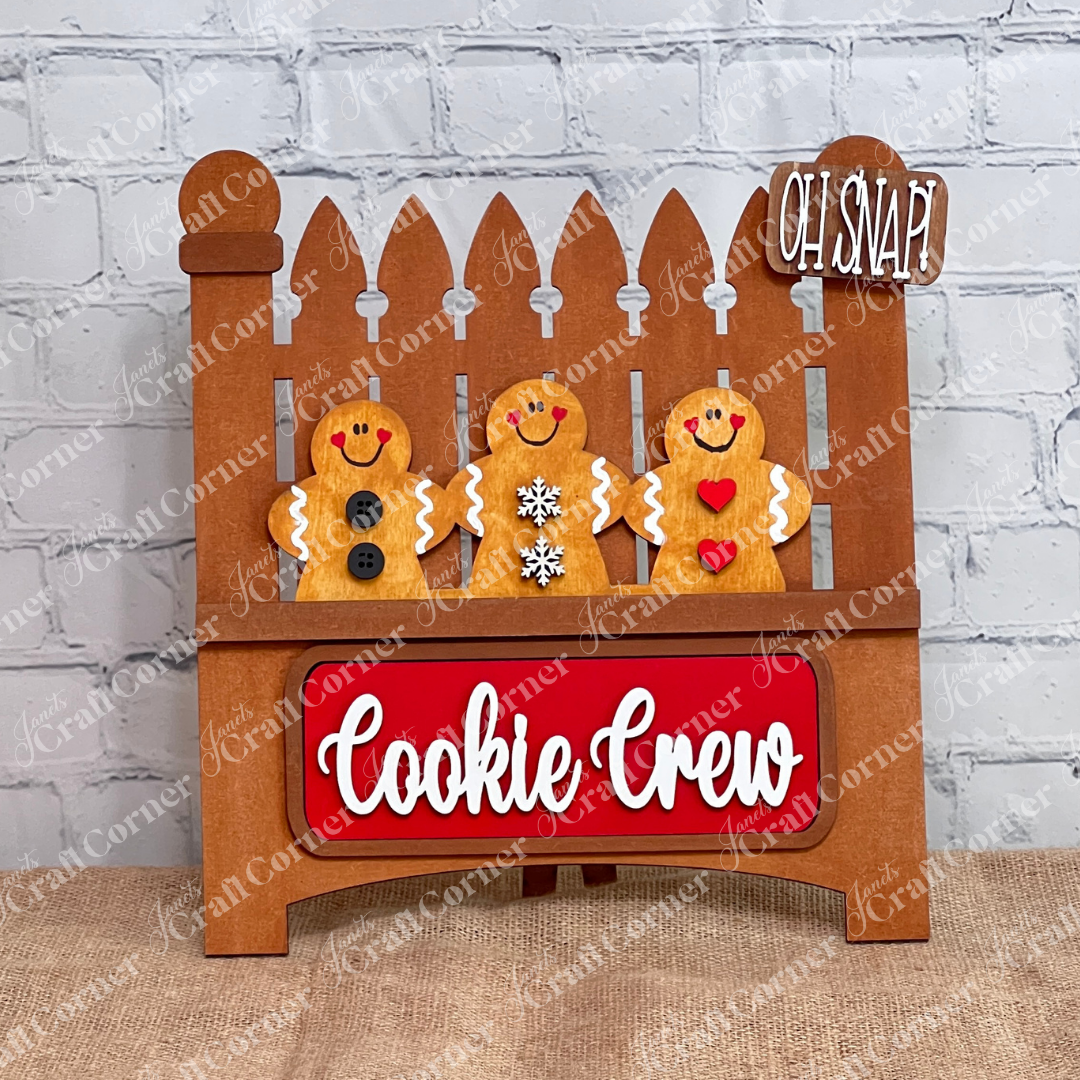 The Cookie Crew Interchangeable Insert by Janet's Craft Corner is a DIY home decor craft kit featuring three uniquely patterned gingerbread men on a white picket fence. The board reads "Oh Snap!" and includes a red "Cookie Crew" sign against a white brick wall, perfect for seasonal inserts.