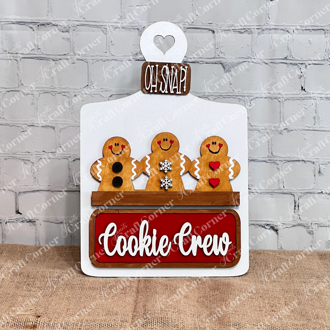 The Cookie Crew Interchangeable Insert by Janet's Craft Corner is a DIY home decor craft kit featuring three uniquely patterned gingerbread men on a white picket fence. The board reads "Oh Snap!" and includes a red "Cookie Crew" sign against a white brick wall, perfect for seasonal inserts.