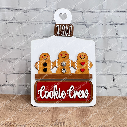 The Cookie Crew Interchangeable Insert by Janet's Craft Corner is a DIY home decor craft kit featuring three uniquely patterned gingerbread men on a white picket fence. The board reads "Oh Snap!" and includes a red "Cookie Crew" sign against a white brick wall, perfect for seasonal inserts.