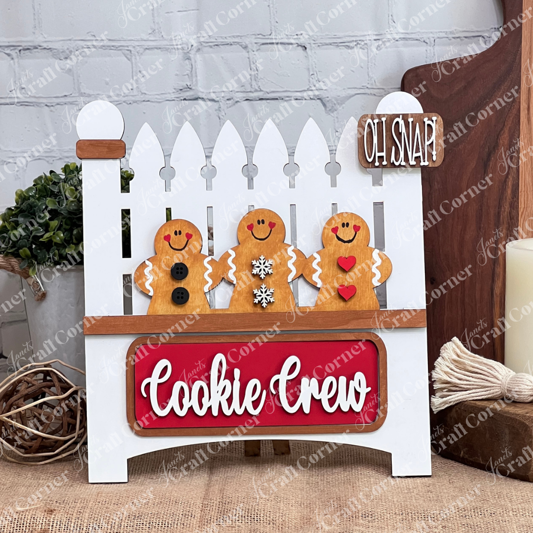 Janet's Craft Corner offers a DIY Garden Fence with Insert kit featuring a charming holiday decoration: a wooden white picket fence with three unique gingerbread figures, a "Cookie Crew" red plaque, and an "Oh Snap!" sign against a brick wall backdrop.