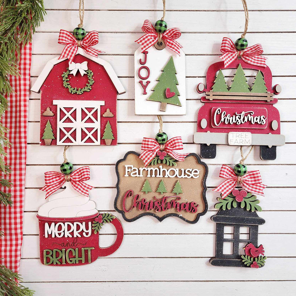 A set of six Country Christmas Ornaments from Janet's Craft Corner adorns a white wall, showcasing a hand-painted DIY craft kit. The collection features charming designs like a red barn, green trees, mistletoe ornaments, a red truck, and a hot chocolate mug. Framed signs with "Joy" and "Farmhouse Christmas" are beautifully accented with red bows and greenery.