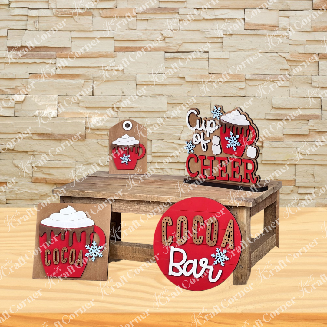 Janet's Craft Corner Cup of Cheer Hot Cocoa Mini Shelf Sitter DIY kit adds cozy winter charm to your home. Featuring cocoa and snowflake designs, this decor sits on a wooden bench with "Cocoa Bar," "Cup of Cheer," and "Cocoa" signs against a stone wall backdrop.