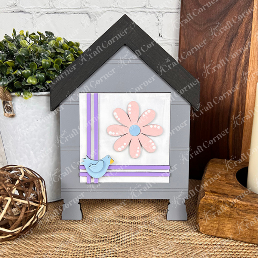 The Bloom & Grow Squares by Janet's Craft Corner is a decorative gray wooden house with a black roof, pink flower, purple ribbons, and blue bird—perfect tiered tray decor for your table alongside plants, a wicker ball, and candle.