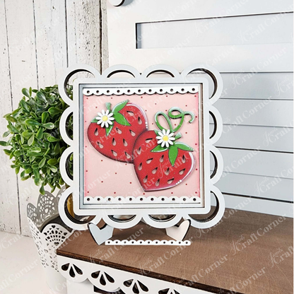 The Strawberry Swappable Squares DIY kit from Janet's Craft Corner features two red strawberries with green stems and white flowers in a scalloped white frame—perfect for tiered tray decor, shown beside a potted plant on a white wooden background.