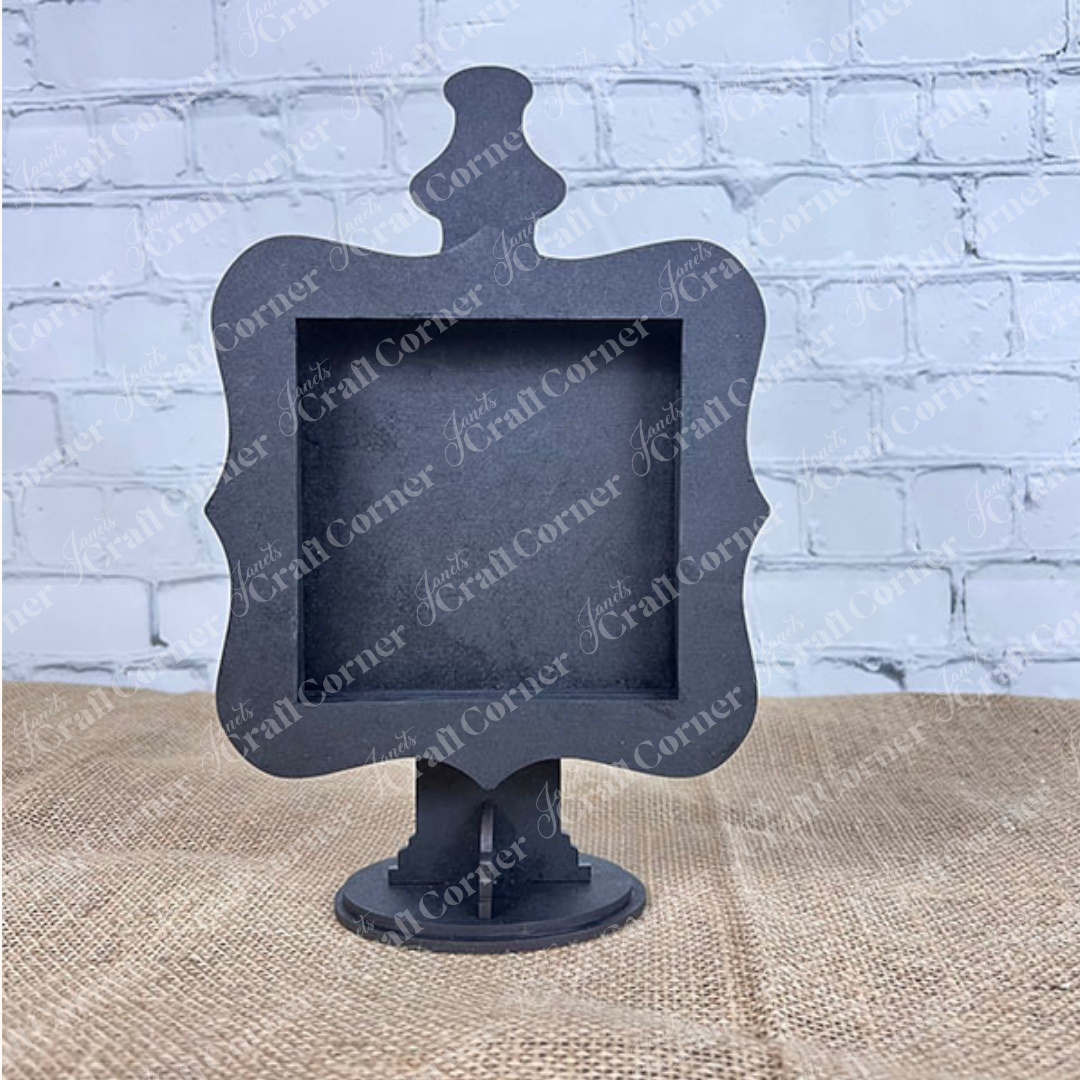 The Heartland Drop In Swappable Squares Holder - DIY Craft Kit (Set of 1) is a black, ornate, square sign holder with a decorative top on a pedestal, perfect for home décor and displayed here on burlap against a white brick wall.
