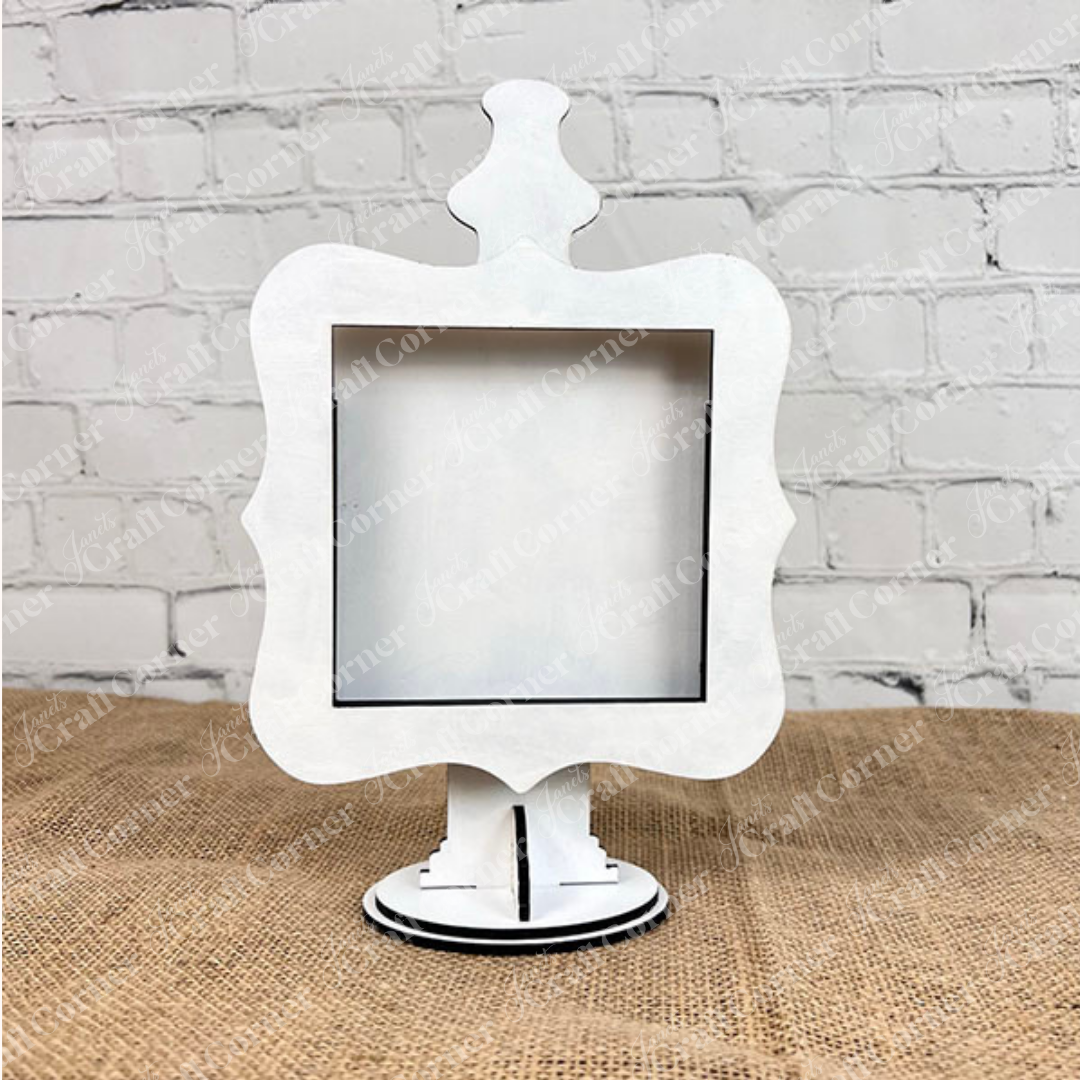 The Heartland Drop In Swappable Squares Holder—an ornate white sign holder with scalloped edges—stands upright on a pedestal, making a charming home décor accent. DIY Craft Kit, Set of 1. Displayed against a white brick wall and burlap surface.