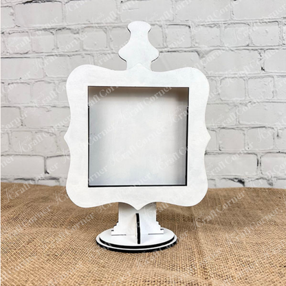 The Heartland Drop In Swappable Squares Holder—an ornate white sign holder with scalloped edges—stands upright on a pedestal, making a charming home décor accent. DIY Craft Kit, Set of 1. Displayed against a white brick wall and burlap surface.