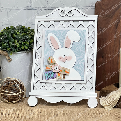 Janet's Craft Corner Cottage Frame for Switchable Farmhouse Signs is a DIY seasonal home decor kit. Display the white frame holding the “HOP” bunny card on a table with plants and rustic accents for a charming farmhouse touch.