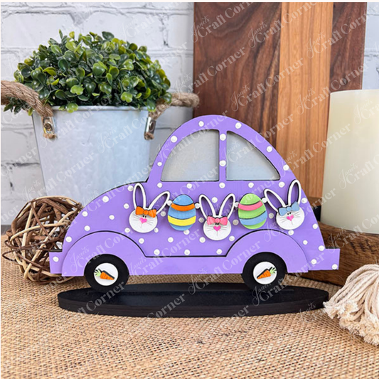 This Easter, the Easter Volkswagen Shelf Sitter from Janet's Craft Corner adds a stylish touch to your home décor. The wooden decoration features a purple car adorned with white polka dots and delightful bunny and egg designs. It is beautifully complemented by a small potted plant, round wicker accents, and a candle in the background.