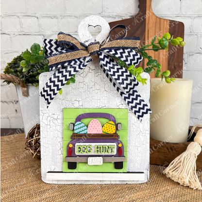 The Hello Easter Squares DIY kit from Janet’s Craft Corner features a cute painted green truck with Easter eggs, "EGG HUNT" text, black-and-white chevron ribbon, greenery accents, and a rustic white background for festive decor.
