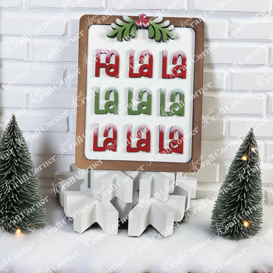 Janet's Craft Corner Fa La La Shelf Sitter DIY kit features "Fa La La La La" in red and green on a white base, surrounded by snow-dusted mini Christmas trees and geometric decor—perfect for cheerful holiday decorating.