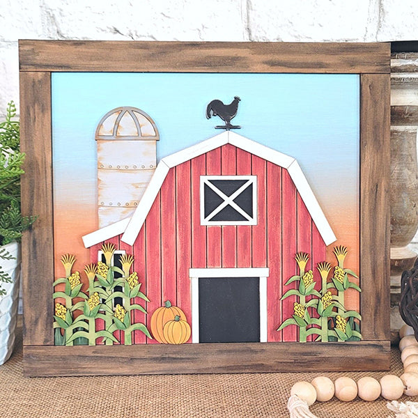 The Fall Barn Scene Leaner from Janet's Craft Corner is a DIY home decor kit that adds rustic charm to any space. This hand-painted framed picture features a red barn with a black door and white trim, complete with a rooster weather vane. The scene includes corn stalks, a pumpkin, and sits beside a silo under a blue and orange sky, making it perfect for enhancing your home's decor.