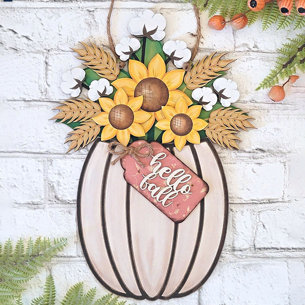 A rustic pumpkin-shaped decoration from Janet's Craft Corner, included in our Fall Floral Pumpkin Door Hanger DIY home decor kit, adorns a white brick wall. It showcases vibrant sunflowers, cotton bolls, and leaves highlighted by a small red tag reading "hello fall," and is beautifully complemented by green ferns and clusters of small orange berries.