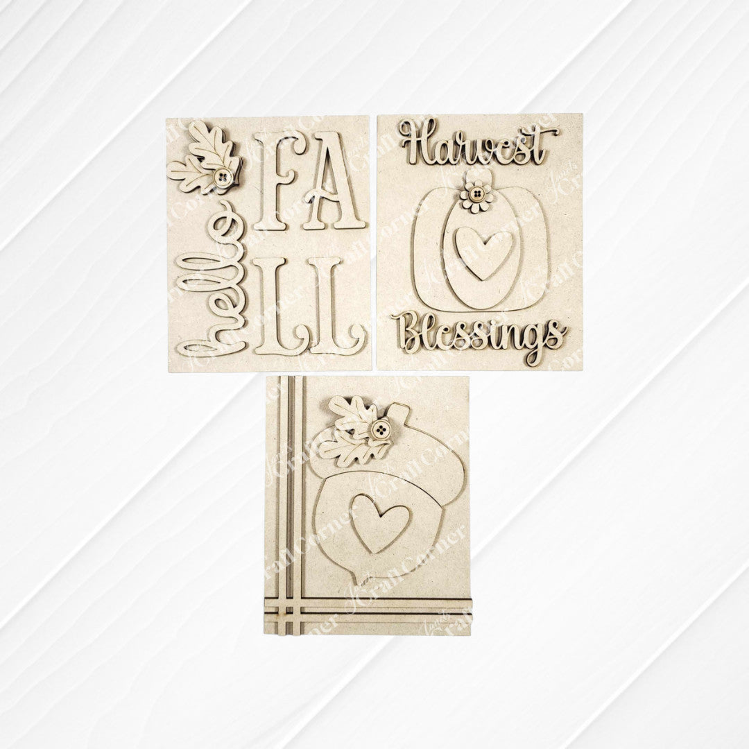 Three Fall Harvest Switchable Farmhouse Signs from Janet's Craft Corner rest on a white wood surface, featuring autumn phrases and images like pumpkins and acorns. This DIY craft kit offers a choice of 3 seasonal home decor signs—perfect for fall decorating!.