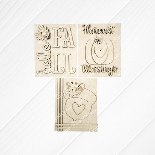 Three Fall Harvest Switchable Farmhouse Signs from Janet's Craft Corner rest on a white wood surface, featuring autumn phrases and images like pumpkins and acorns. This DIY craft kit offers a choice of 3 seasonal home decor signs—perfect for fall decorating!.