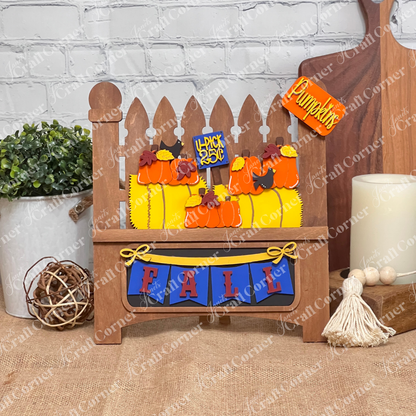 Janet's Craft Corner presents a delightful DIY home decor kit featuring a Garden Fence with an insert. It includes a wooden picket fence decorated with paper pumpkins, a "Pumpkins" sign, and "4 Pack 25¢" label, completed by a blue and red "FALL" banner set against a rustic white brick wall.