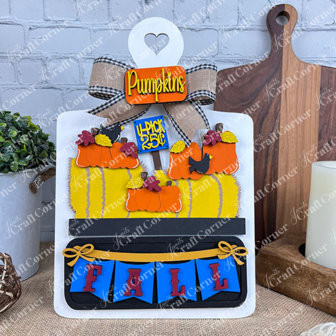 A decorative fall-themed display includes the Fall Hay Bale Interchangeable Insert - Painted from Janet's Craft Corner. It features a small board with a heart cutout, adorned with hand-painted ornaments and a "Pumpkins" sign. Surrounded by pumpkins, a yellow plaid backdrop, and truck, it spells out "FALL" in blue with red letters against a brick wall, offering interchangeable fall décor options.
