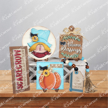 A display of fall-themed wooden decor from Janet's Craft Corner, featuring the Fall Scarecrow Mini Shelf Sitters DIY craft kit, “Autumn Blessings” sign, mini pumpkin shelf sitter, and a beaded tassel with autumn tags on a white bench.