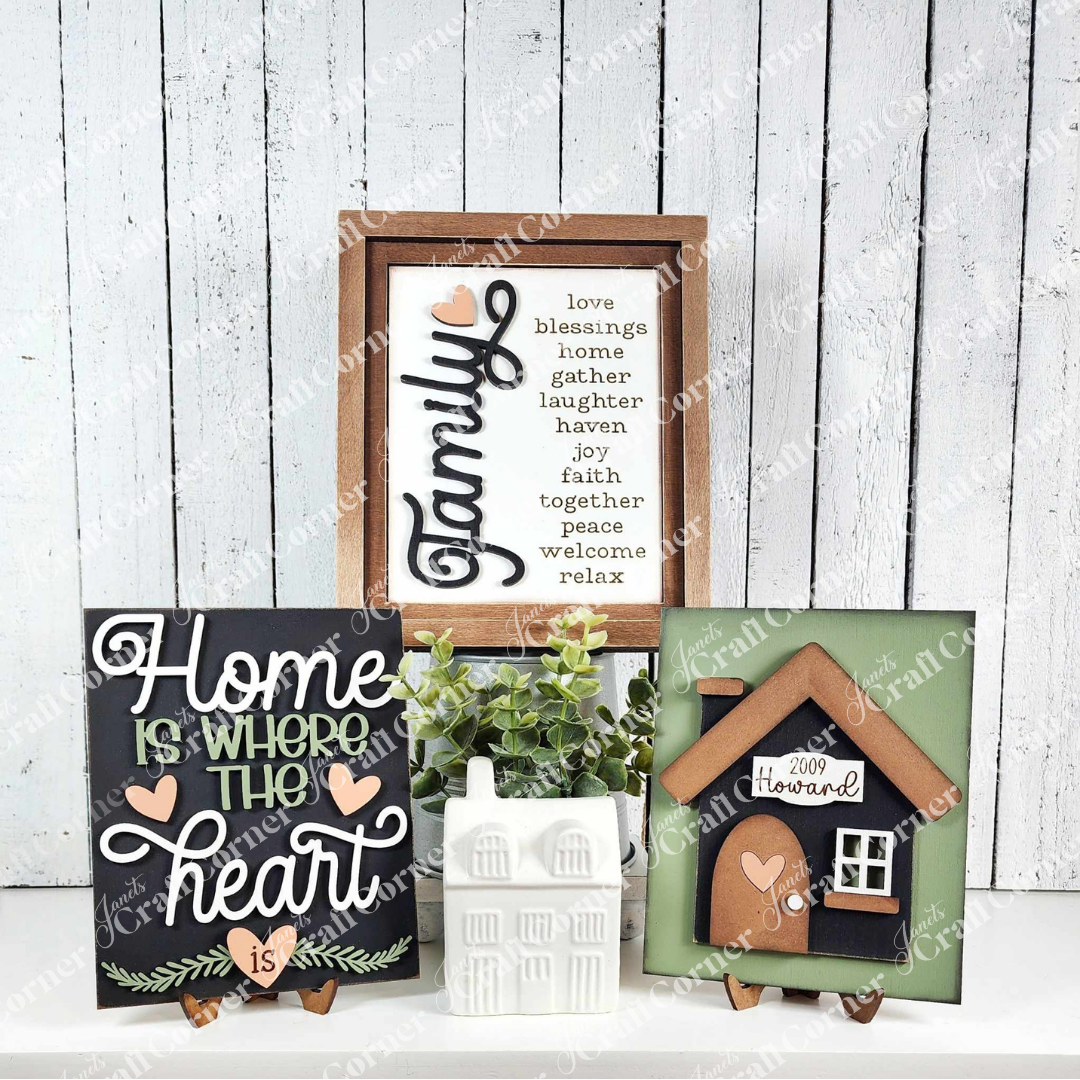 Displayed against a white plank wall, signs from Janet's Craft Corner exude rustic charm. One reads "Family," featuring words like "love" and "blessings," while another says, "Home is where the heart is." A house farmhouse sign completes these cozy Family-Home Interchangeable kits.