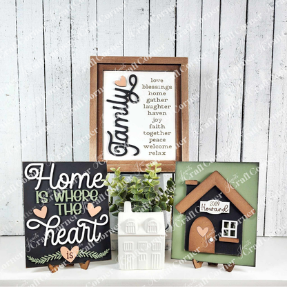 Displayed against a white plank wall, signs from Janet's Craft Corner exude rustic charm. One reads "Family," featuring words like "love" and "blessings," while another says, "Home is where the heart is." A house farmhouse sign completes these cozy Family-Home Interchangeable kits.