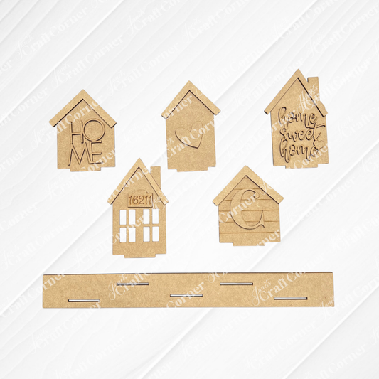 Five small, house-shaped wooden pieces and one long rectangular base from the Janet’s Craft Corner Family Houses Shelf Sitter DIY home decor craft kit (set of 1) are arranged on a white surface. Ideal for personalized home accents or crafting, two houses feature engraved messages: “HOME” and “home sweet home.”.