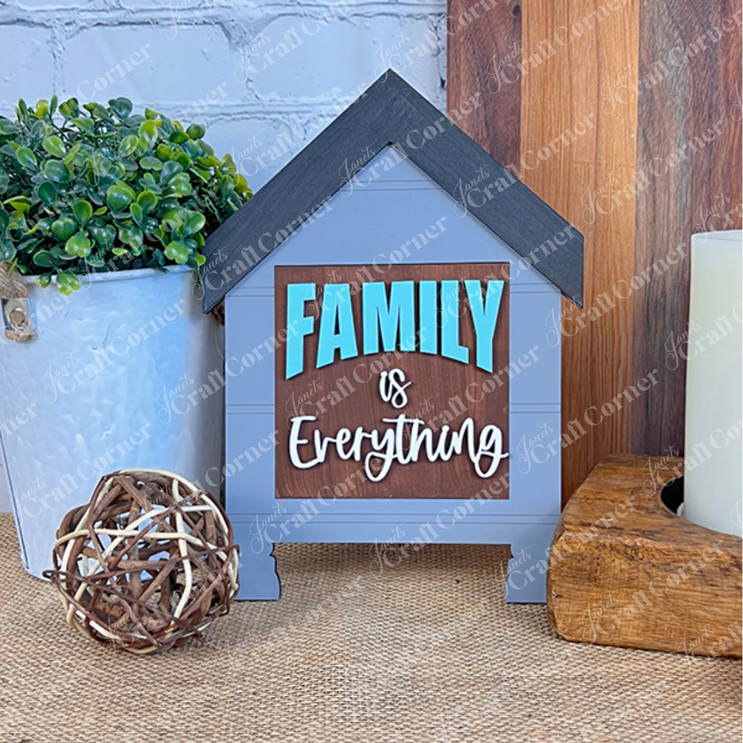 Janet's Craft Corner’s Family Home Squares features a house-shaped sign reading “Family is Everything” in blue and white, arranged with a green potted plant, woven ball, and wood cut-outs for stylish tiered tray decor.