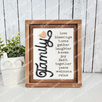 The painted sample of the Family Words Farmhouse Sign by Janet's Craft Corner features "Family" prominently in cursive, surrounded by words like love, blessings, laughter, and peace. It pairs beautifully with a white ceramic house and green plant on whitewashed wood panels.